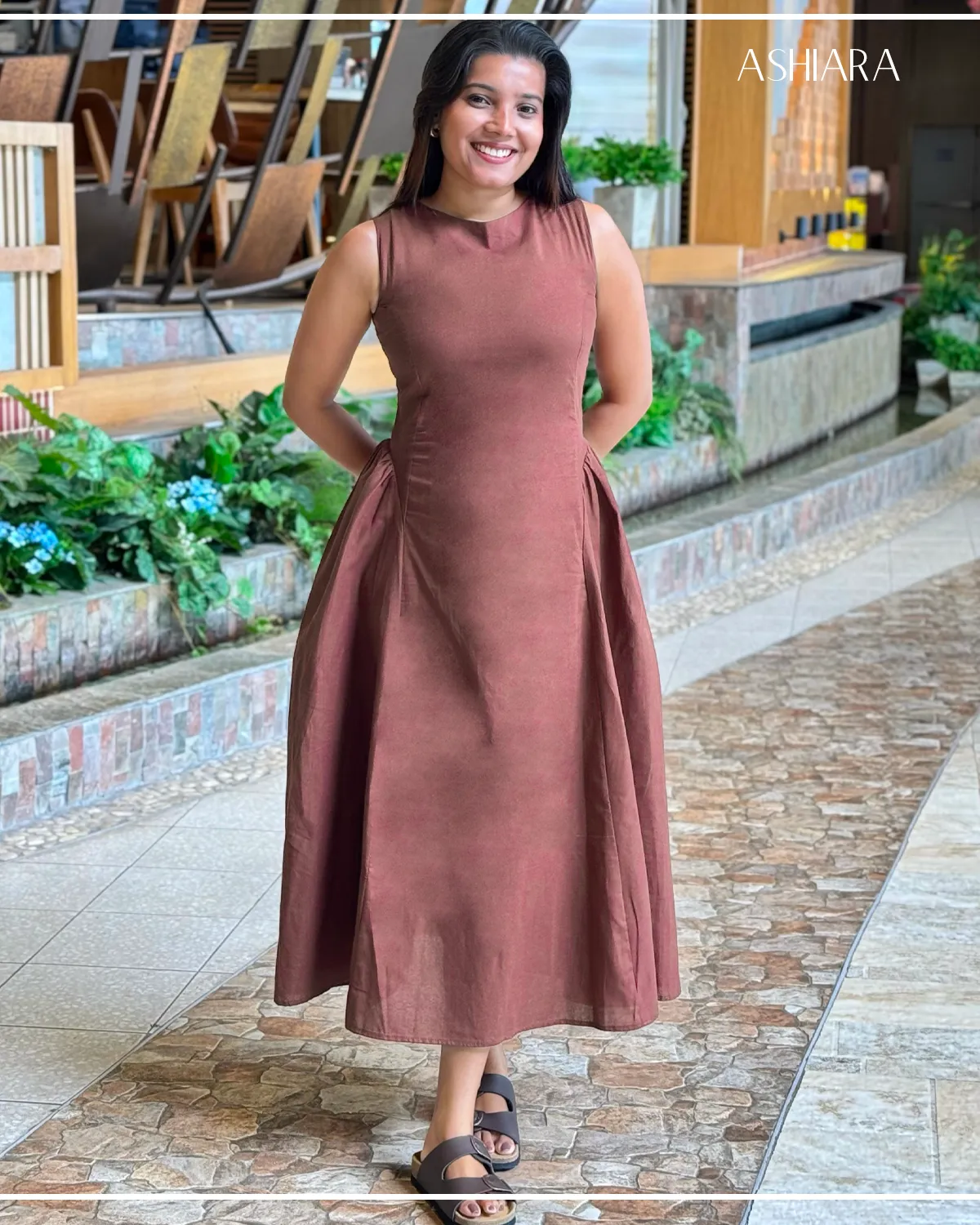 Full frontal view of a sleeveless, brown midi dress with a round neckline. Worn by a model standing on tiled pavement with garden decor. Label 'ASHARA' visible at the top right of the image.