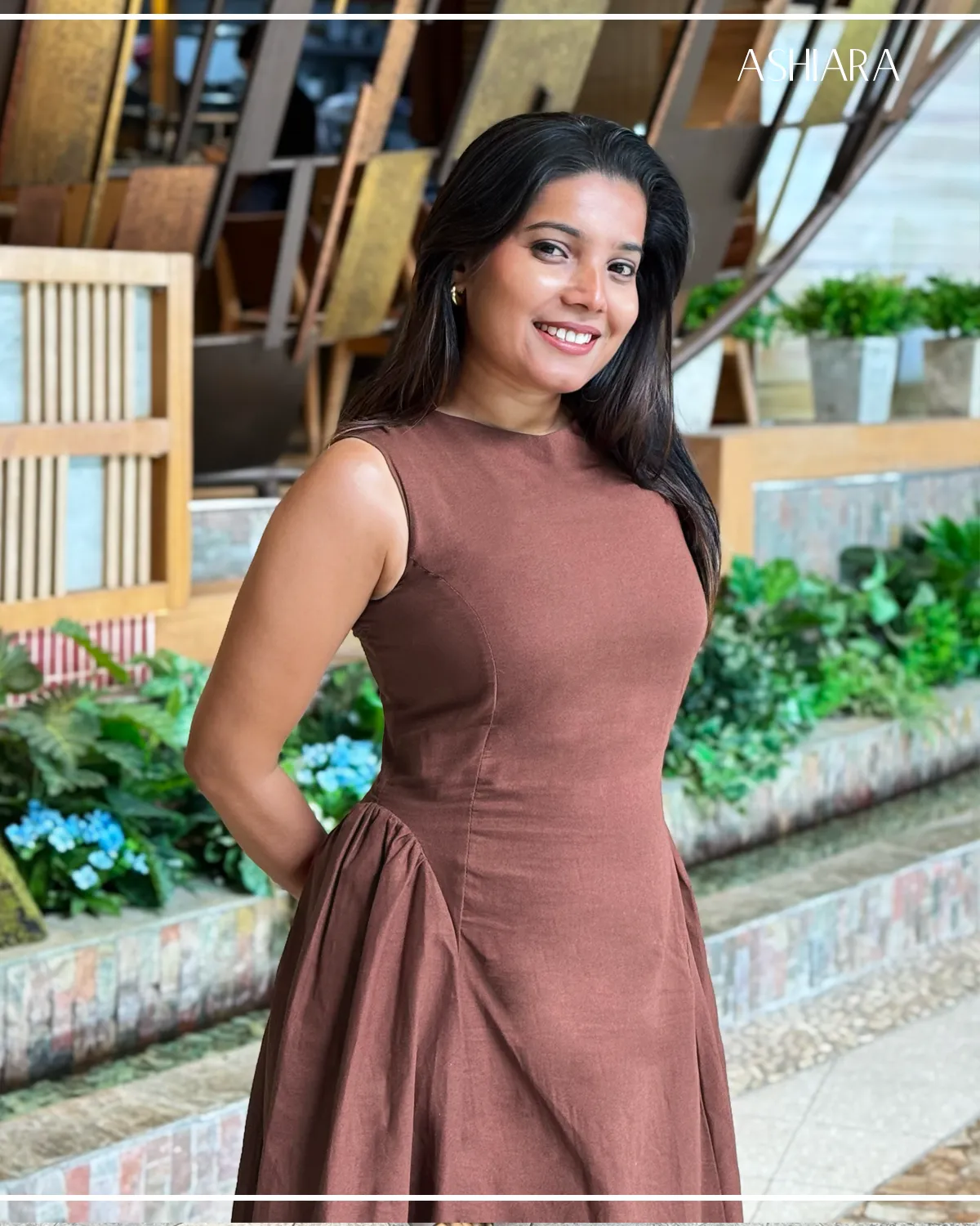 A brown, sleeveless dress with a fitted upper and flared skirt. Worn by a model against a backdrop of plants and artistic installations. Branding 'ASHARA' visible in the top right corner.