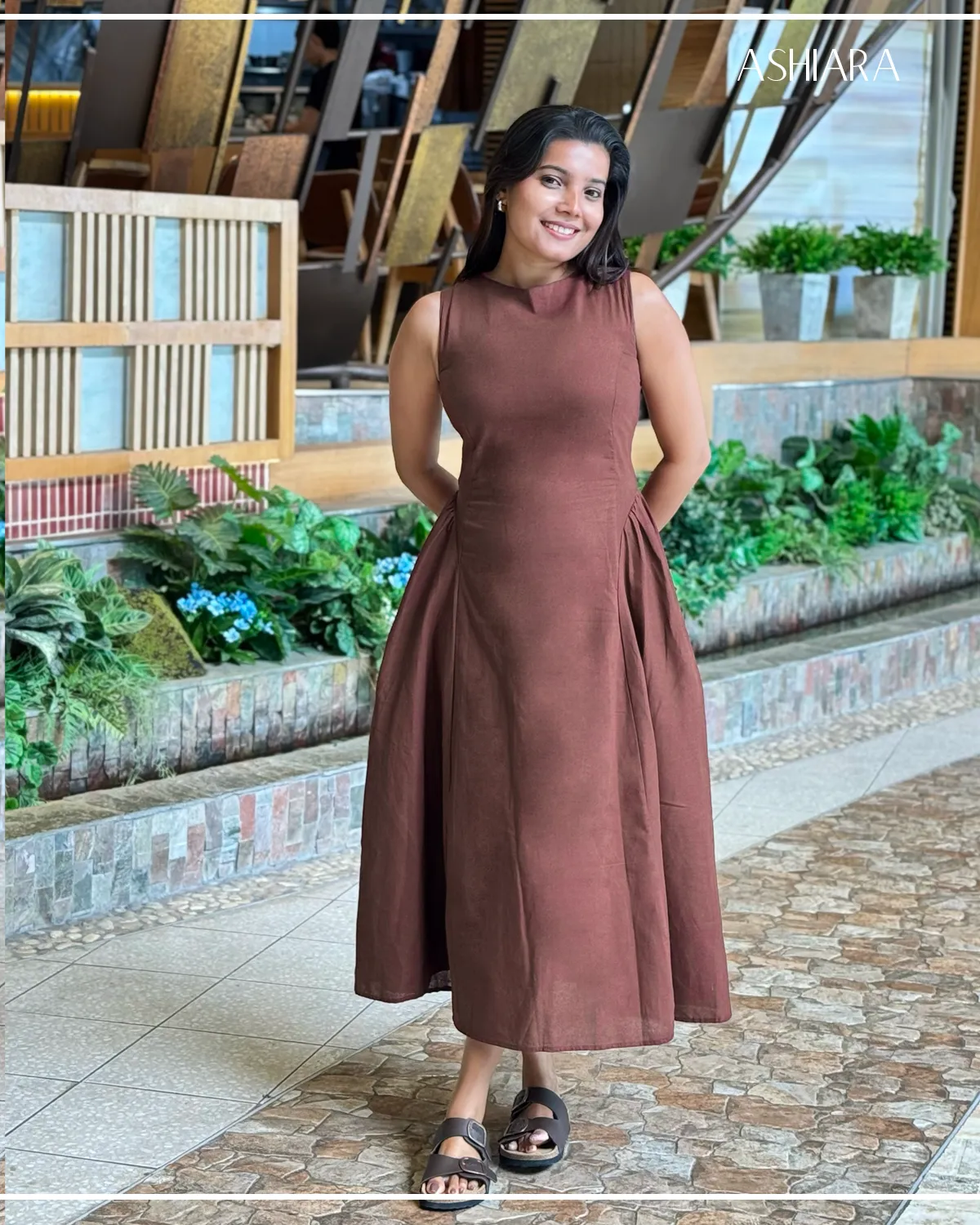 A sleeveless, brown, ankle-length dress with a fitted bodice and flared skirt. It's worn by a model standing on a stone floor in a garden-like setting. Branding 'ASHARA' visible at the top right.